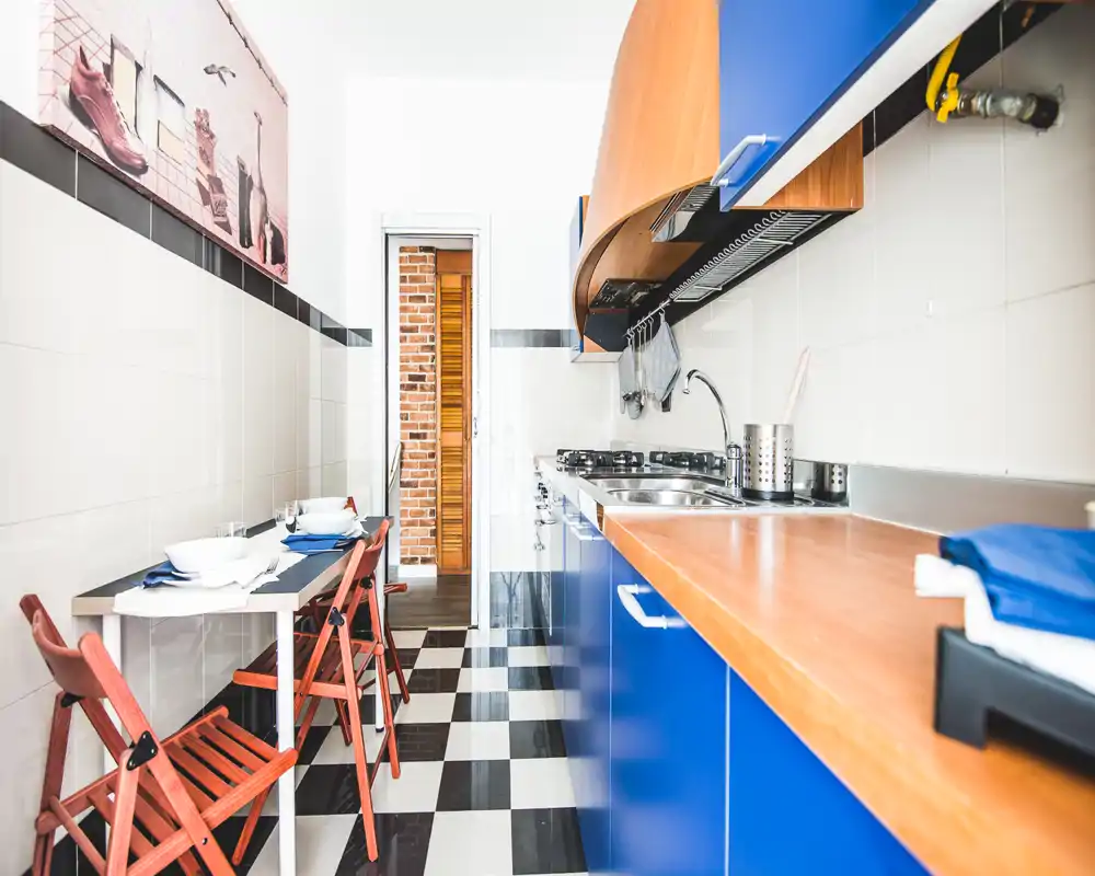 Cocina estrecha y luminosa con muebles azules, encimera de madera y una pequeña zona de comedor con sillas plegables; estilo moderno y suelo en damero blanco y negro.