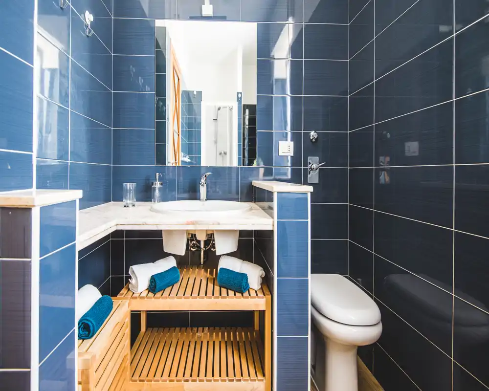 Bagno moderno con piastrelle lucide blu scuro, lavabo bianco incassato in un piano in marmo, scaffalatura in legno con asciugamani arrotolati e WC a destra — luminoso, pulito e allestito con cura per affitti brevi.