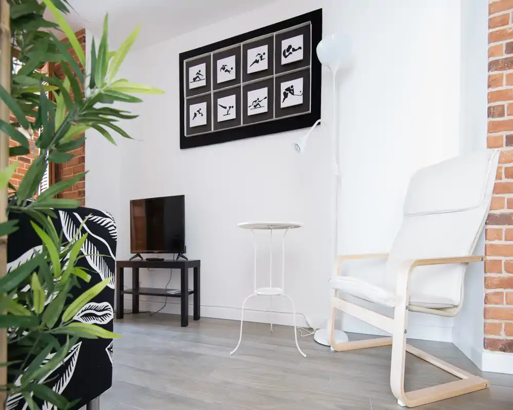 Zona de estar moderna y luminosa con un sillón cómodo, una pequeña TV sobre un mueble y cuadros decorativos: un rincón de salón compacto y bien decorado, ideal para relajarse.