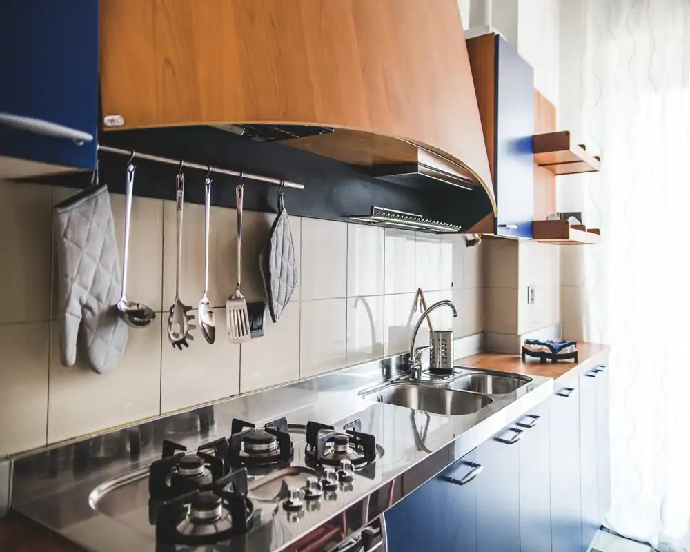 Cocina moderna y luminosa con encimera de acero inoxidable, cocina de gas y fregadero doble; muebles de madera y utensilios colgados aportan calidez y orden.