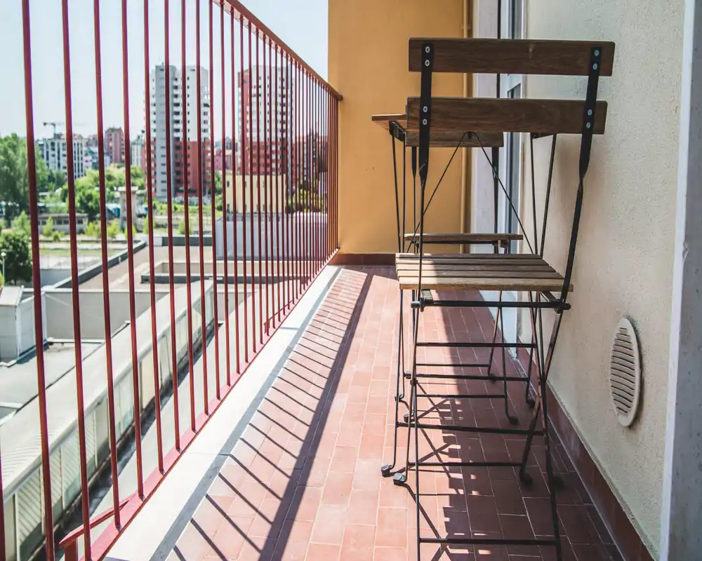 Balcón soleado con suelo de baldosa, barandilla metálica y una pequeña mesa y silla plegables de madera — ideal para un café por la mañana con vistas urbanas.