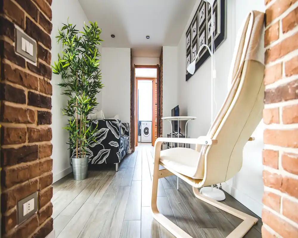 Zona de estar compacta y con estilo con sofá estampado, butaca de madera clara, planta y ladrillo visto; luz natural desde la puerta del fondo.