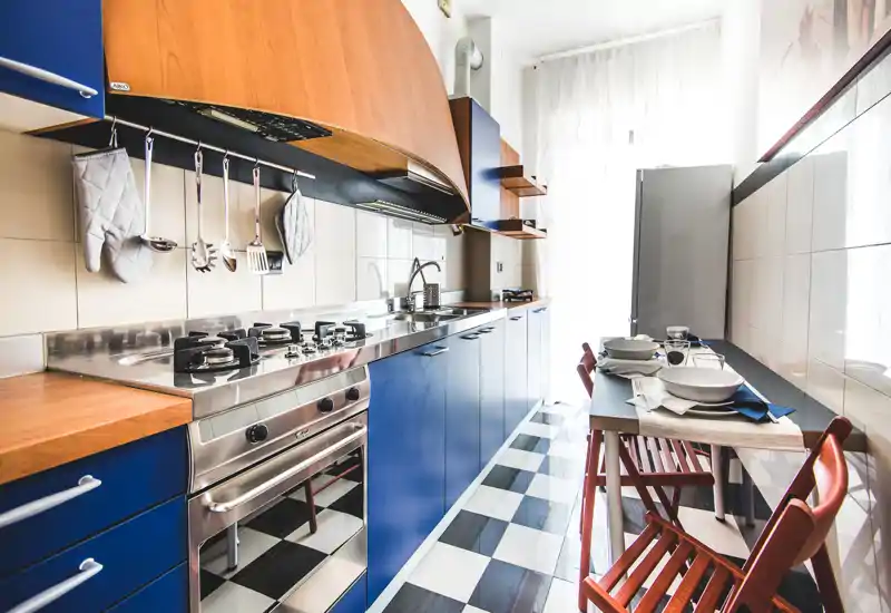 Cucina a corridoio luminosa e moderna con mobili blu, elettrodomestici in acciaio inox e un piccolo angolo pranzo per due. La luce naturale e il pavimento a scacchi rendono lo spazio accogliente per cucinare e consumare pasti informali.