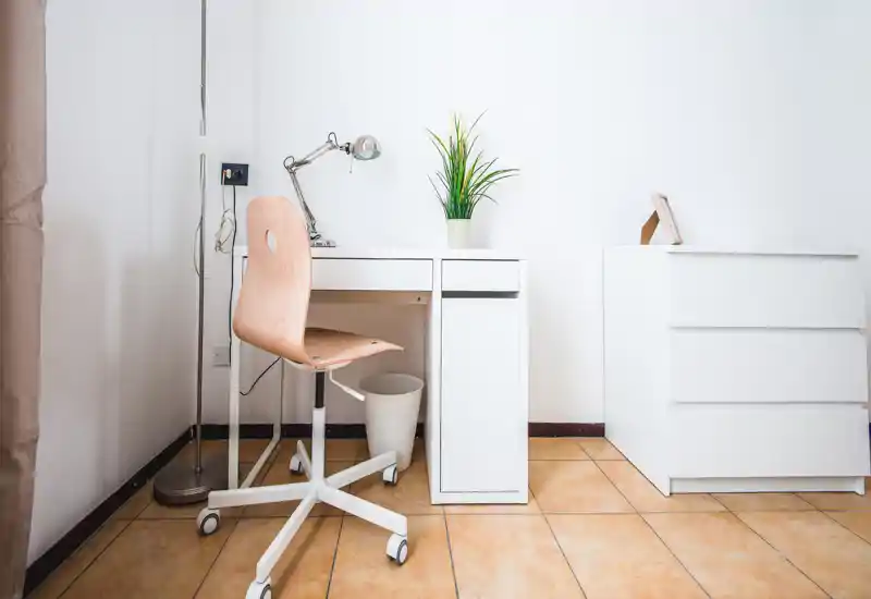 Minimal bedroom corner with a white desk, wooden swivel chair and a small chest of drawers; bright, clean and simply styled with a potted plant accent.