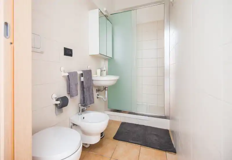 Compact, modern bathroom with a shower enclosure, wall-mounted sink, bidet and toilet. Neutral tiles and coordinated gray towels create a clean, minimal look.