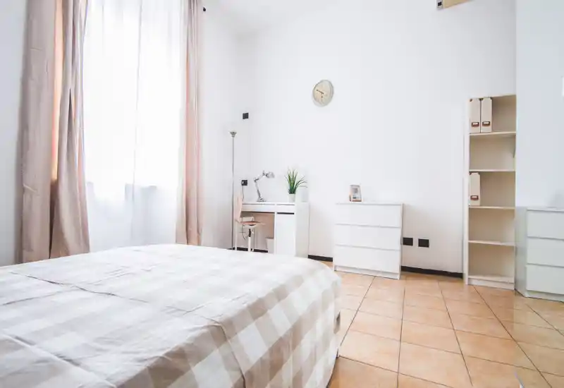 Bright, minimal bedroom with a large bed dressed in neutral checked linens, a small desk, dresser and shelving; generous natural light from a tall window creates an airy atmosphere.