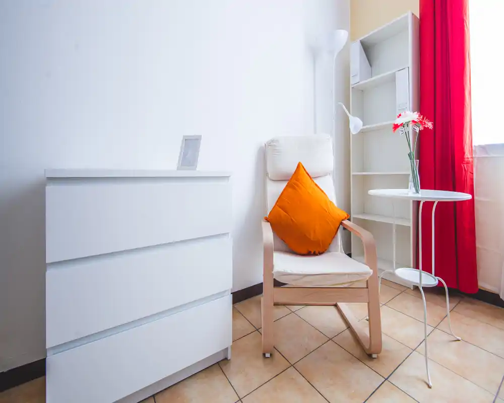 Bright, minimal bedroom corner with a comfortable armchair, orange cushion, white chest of drawers and a small round table by the window.