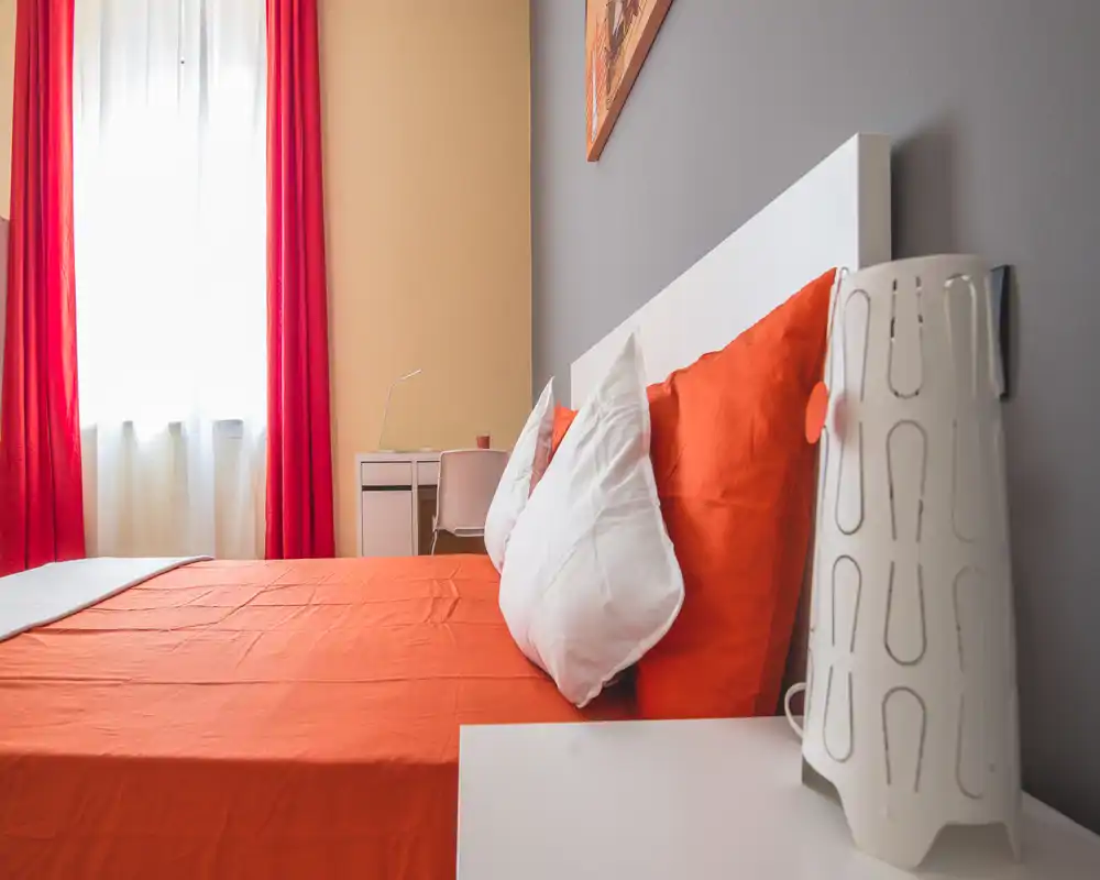 Bright, modern bedroom with orange linens and white headboard; natural light from a large window and coordinated pillows create a warm, inviting atmosphere.