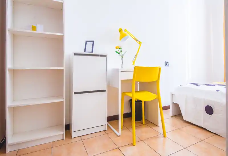Bright, minimal bedroom corner with a small desk, yellow chair and a partially visible bed. Clean white shelving and modern accents create a fresh, airy feel ideal for single occupants or student rentals.
