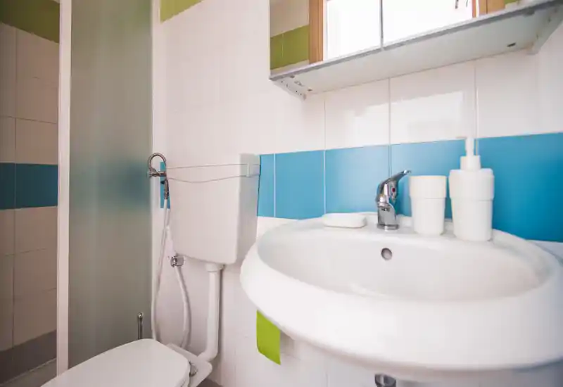 Clean, compact bathroom with a white sink, chrome faucet, soap dispenser and visible toilet; tiled walls feature blue and green accents and a frosted glass shower door.