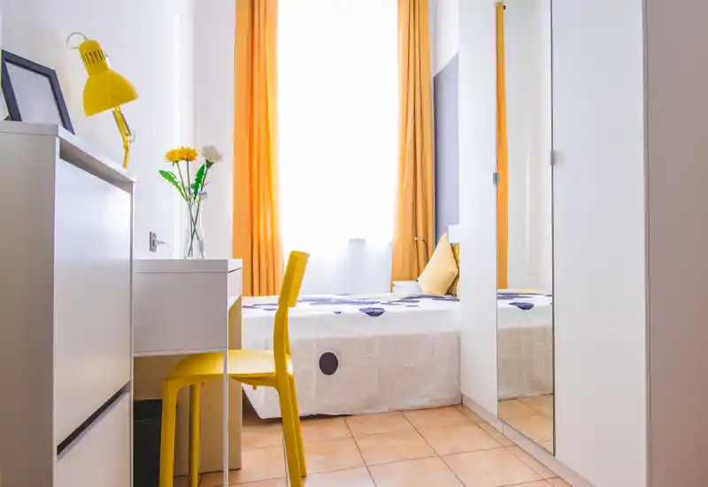 Bright, compact bedroom with a single bed, warm orange curtains and coordinated yellow accents. A desk and chair sit beside the bed, and a mirrored wardrobe adds depth and light.