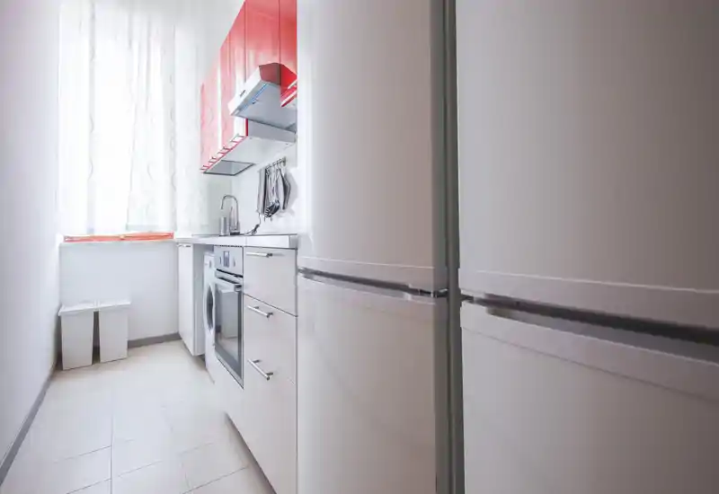 Bright, narrow modern kitchen with white cabinetry, integrated oven and washing machine, and a large refrigerator. Natural light from the window highlights clean surfaces and red accents for a fresh look.