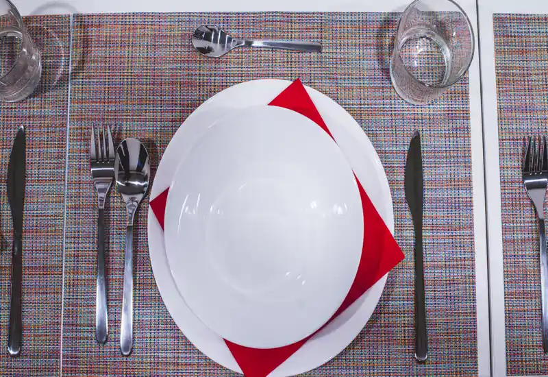 Close-up of a neatly arranged dining place setting with white plates, a red napkin, cutlery and a water glass on a woven placemat.
