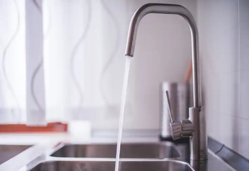 Modern stainless-steel kitchen sink and faucet with running water, softly lit by natural light from a nearby window — clean and minimalist look ideal for listing close-up kitchen details.