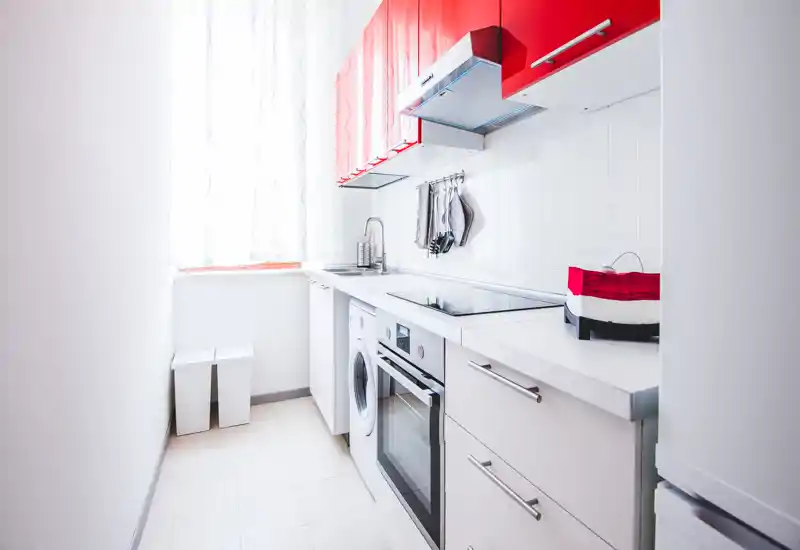 Bright, modern galley kitchen with red upper cabinets, integrated oven and induction hob, plus a washing machine and tidy countertop — ideal for compact urban apartments.