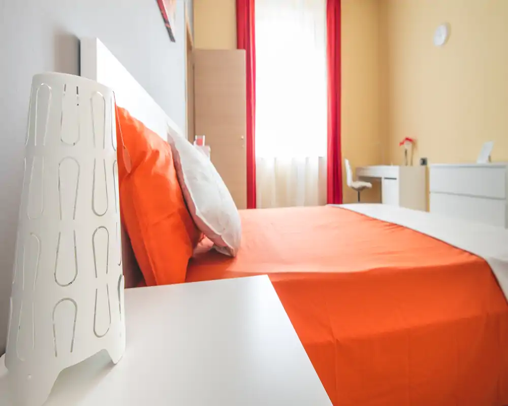 Bright, modern bedroom with a large orange-dressed bed, white bedside table and natural light from a window. Clean, minimal furnishings and a small desk area make this room ideal for comfortable stays.