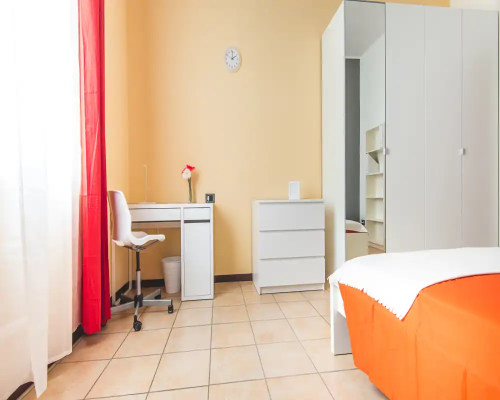 Bright, tidy bedroom with an orange bedcover, white furniture and a small desk by the window. Natural light and clean lines give a fresh, welcoming feel.