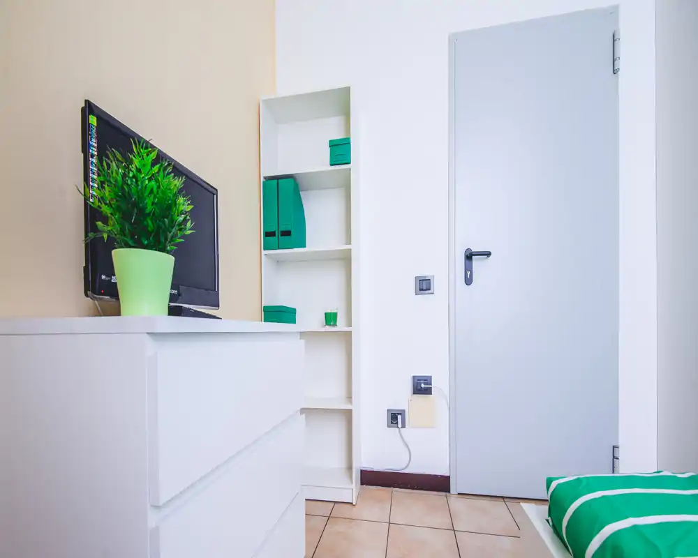 Bright, minimal bedroom corner with a small visible bed, white chest of drawers, shelving and a TV; green accents create a fresh, modern look.