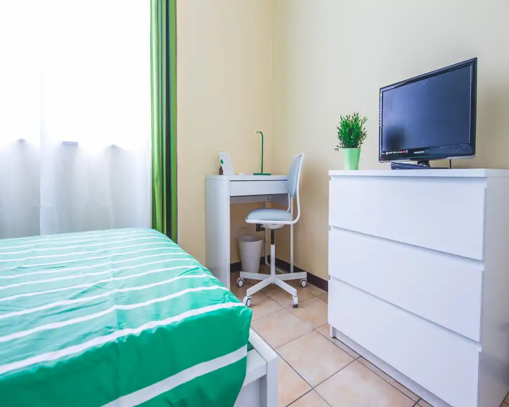 Bright, tidy single bedroom with a green-striped bedspread, white dresser and small desk — simple, well-lit space ideal for a single tenant.