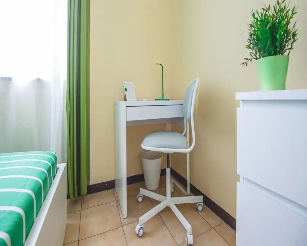 Bright, compact bedroom corner with a single bed, a small white desk and a rolling chair; green accents and a potted plant create a fresh, tidy atmosphere.