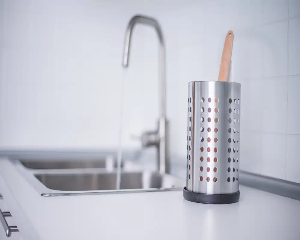 Bright, minimal kitchen countertop with a stainless-steel utensil holder and a double sink with running water — clean, modern styling ideal for rental listing photos.