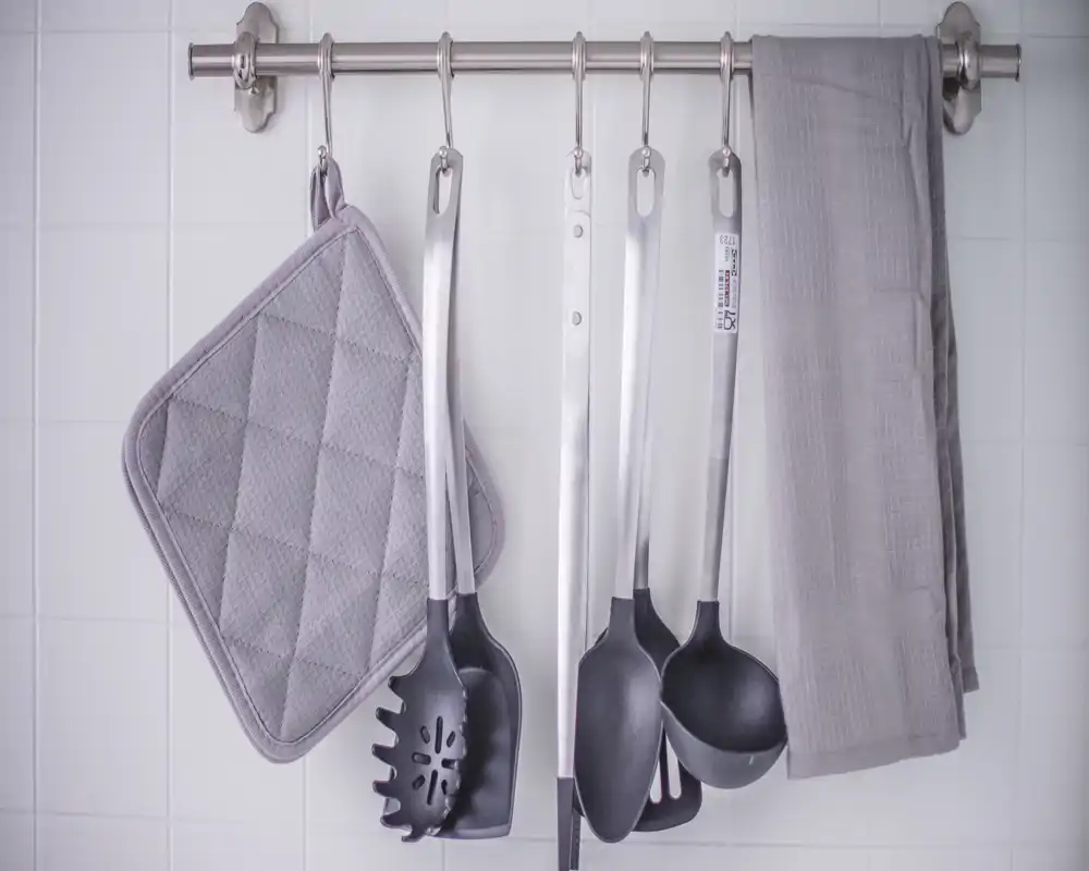 Close-up of kitchen utensils and textiles hanging on a wall rail over tiled backsplash; neat, well-lit and styled for rental photo use.