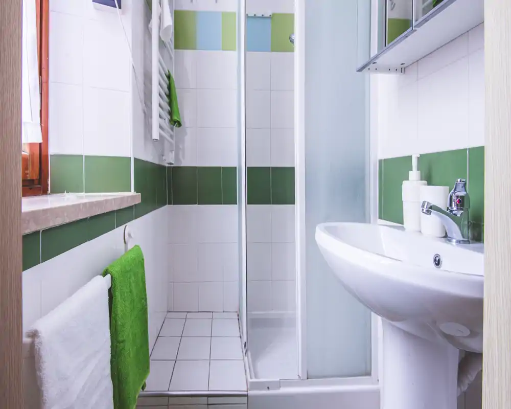 Compact, well-lit bathroom with a corner shower, white sink and green-accented tiles. Clean, simple fixtures and fresh towels create a practical space for guests.