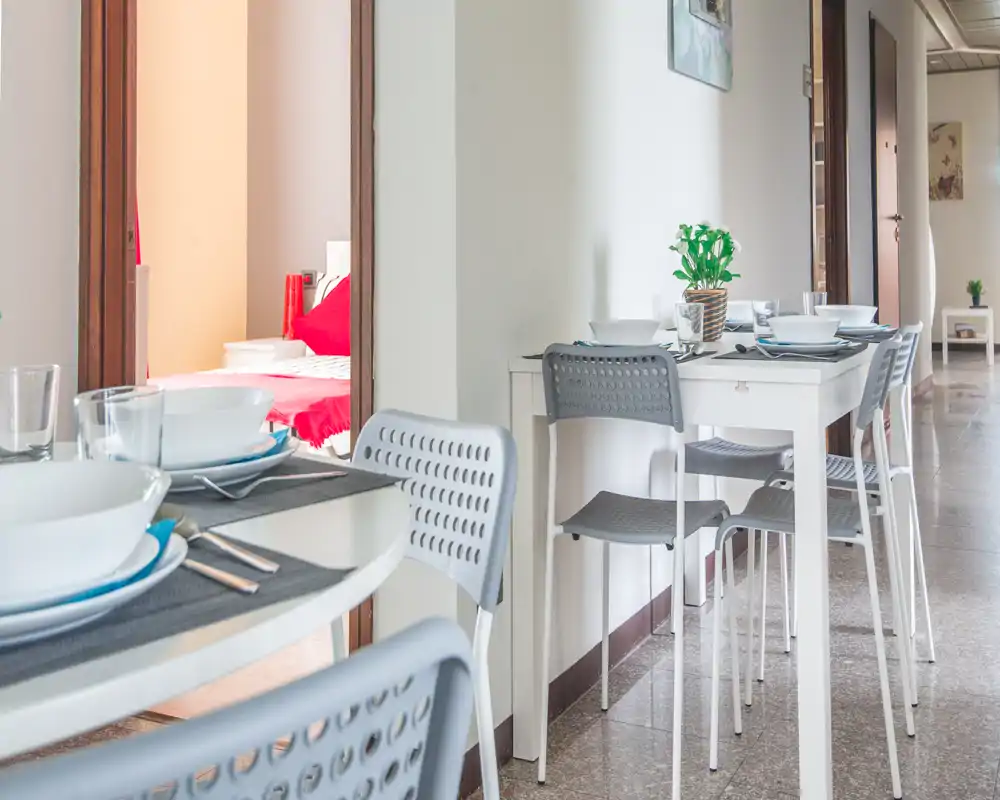 Well-staged dining area with modern white tables and grey chairs, set with plates and glassware; bright, tidy space adjacent to a bedroom visible through an open door.