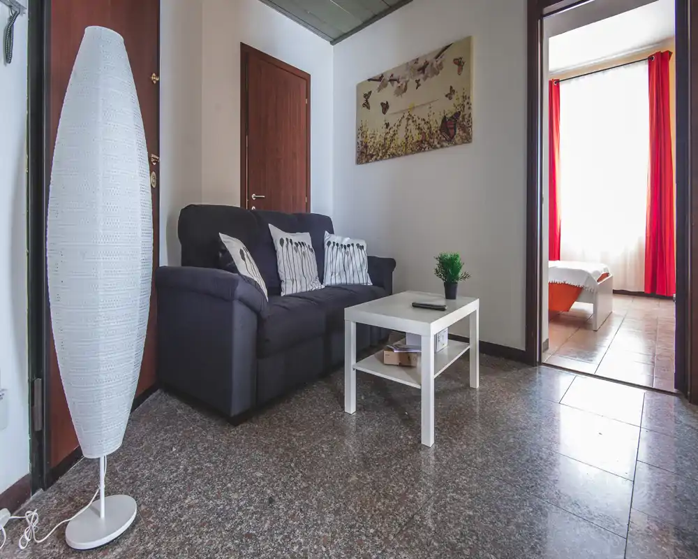 Compact living area with a dark two-seater sofa, white coffee table and decorative cushions; bright doorway leads to a bedroom with red curtains.