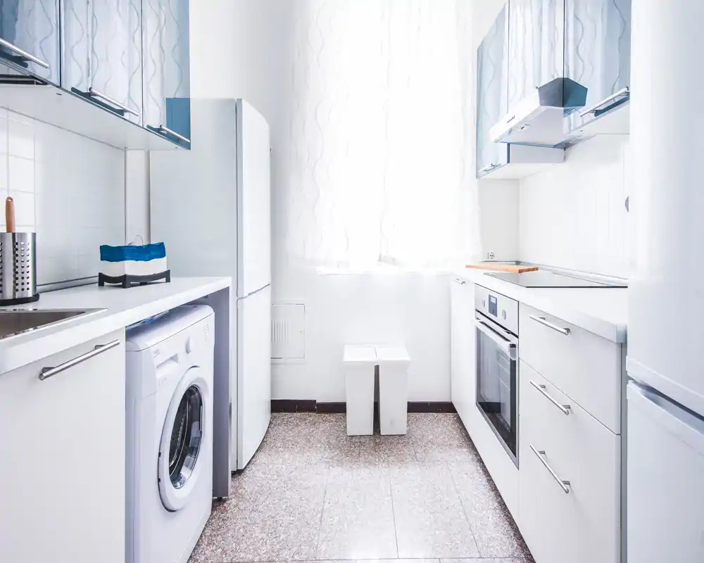 Bright, compact modern kitchen with white cabinetry, built-in oven, fridge and a washing machine; clean minimalist style and natural light from a large window.