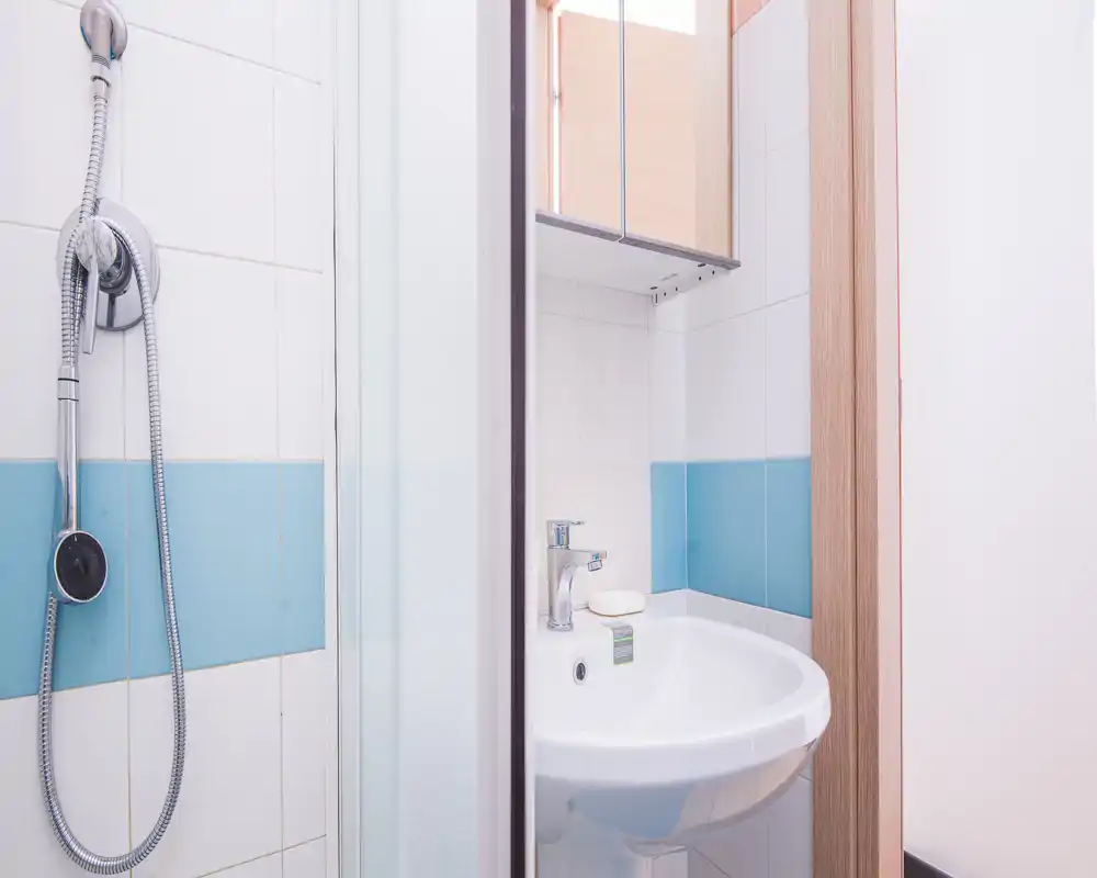 Compact, bright bathroom with a wall-mounted sink, mirrored cabinet and a shower with handheld head—clean white tiles with a blue accent strip.