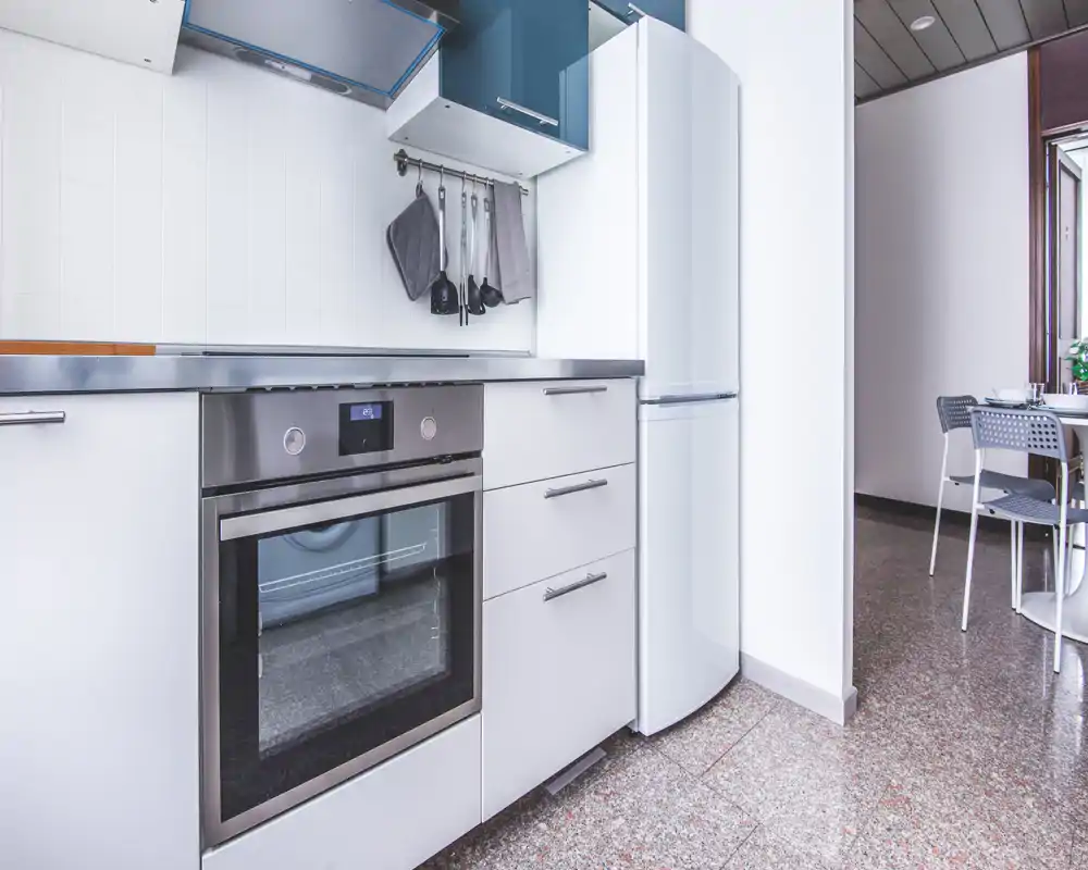 Modern, compact kitchen with stainless-steel oven, white cabinets and a tall refrigerator; clean surfaces and hanging utensils create a practical cooking area.