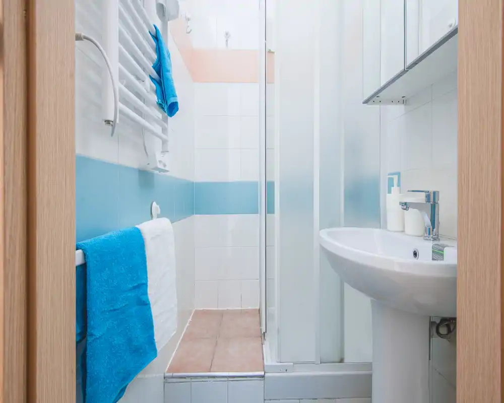 Bright, compact bathroom with a pedestal sink, glass shower enclosure and towel rails; white tiles with blue accent stripe and fresh towels.