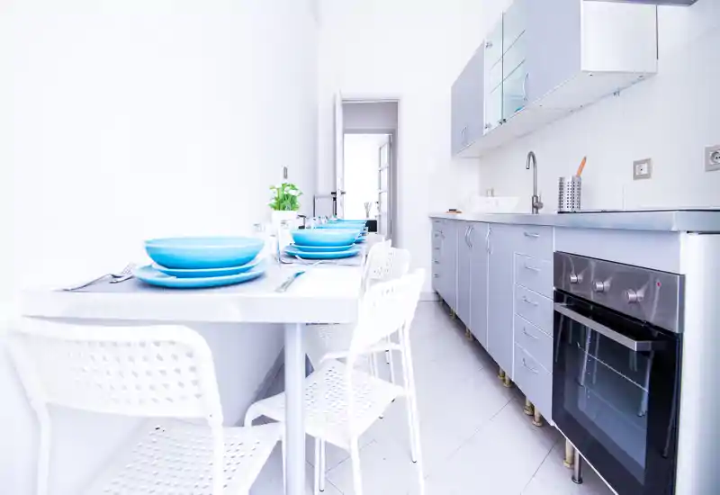 Bright modern galley kitchen with stainless oven, sink and a small dining table set with blue dishware; minimalist, well-lit space ideal for photos of compact apartments.