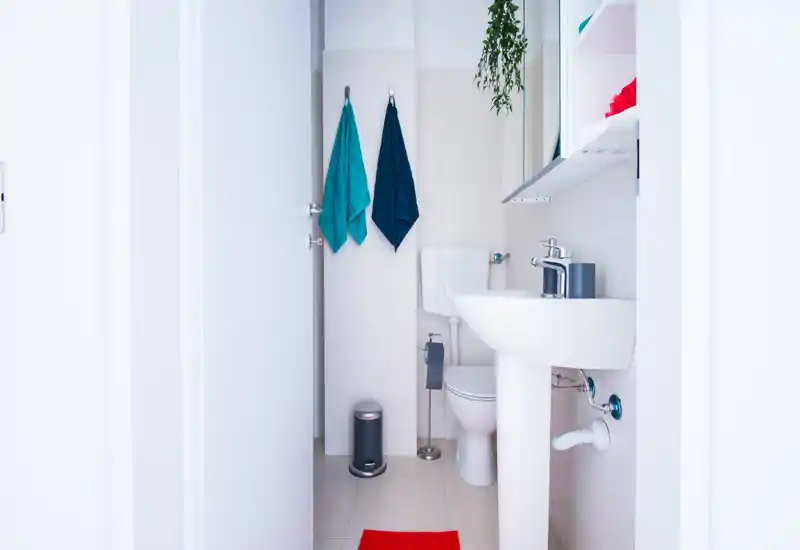 Compact, modern bathroom with a pedestal sink, toilet, towel hooks and bright white tiles; neat accessories and a red bath mat add color to the clean design.