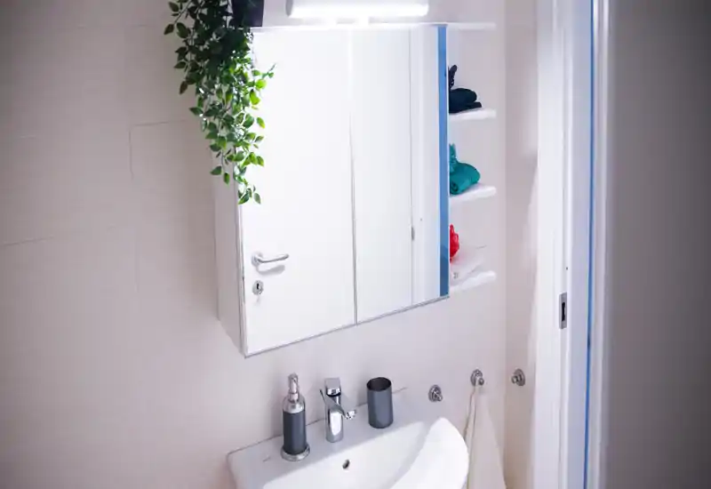 Bright, modern bathroom sink area with a mirrored medicine cabinet, faucet, soap dispenser and small shelves holding folded towels.