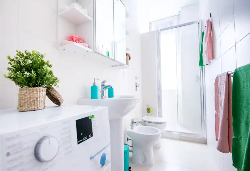 Bright, modern bathroom with a shower cabin, sink, bidet and washing machine. The space is well lit and neatly staged with colorful towels and accessories.