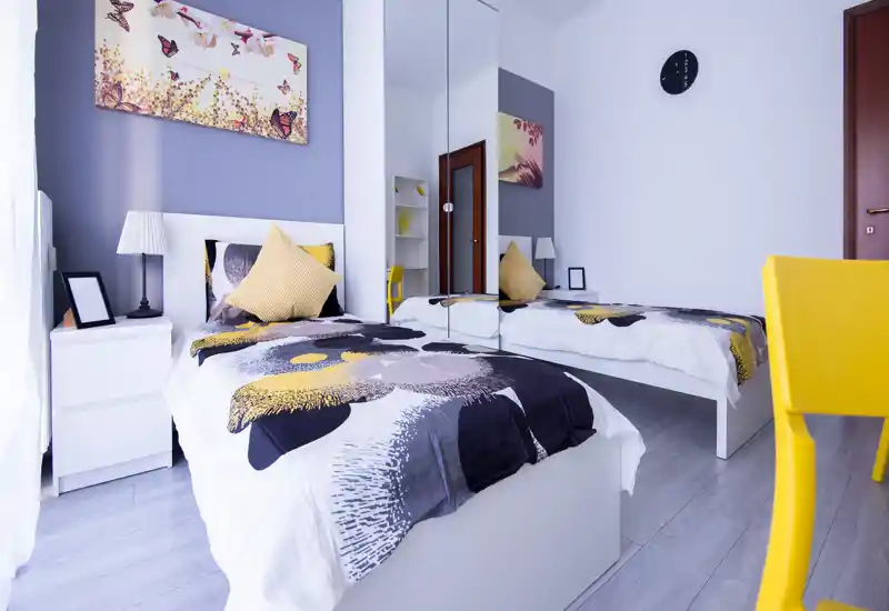 Habitación moderna con dos camas individuales bien hechas, ropa de cama coordinada y toques amarillos alegres. El armario con espejo y la luz natural hacen que la estancia parezca amplia y fresca.