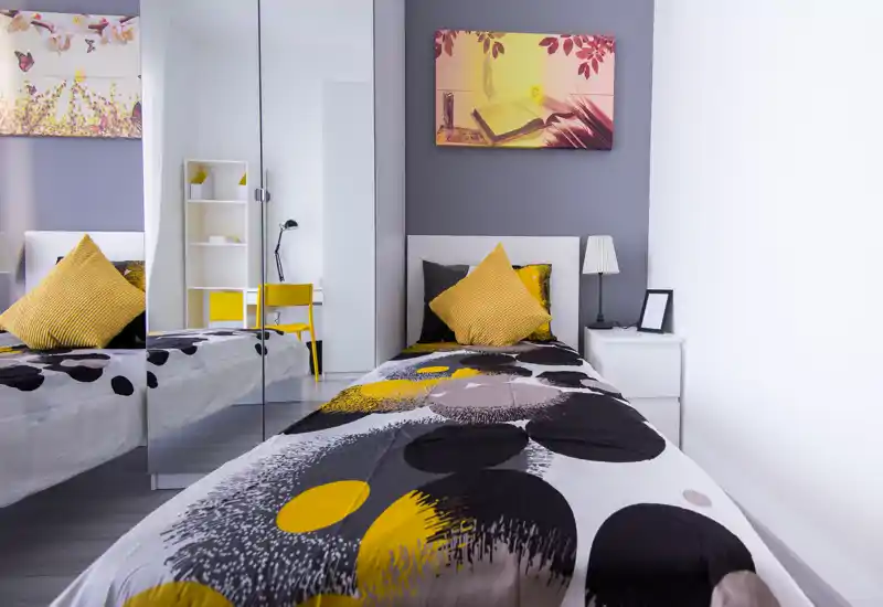 Dormitorio individual moderno y luminoso con ropa de cama estampada, cojines amarillos y armario con puertas espejo que aporta profundidad. Mesita con lámpara y cuadro en la pared, acogedor y perfecto para un anuncio.