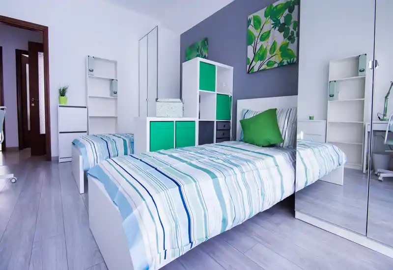 Dormitorio moderno y luminoso con una cama individual bien arreglada, acentos verdes, estanterías y armario con espejo que crean una atmósfera fresca y aireada.