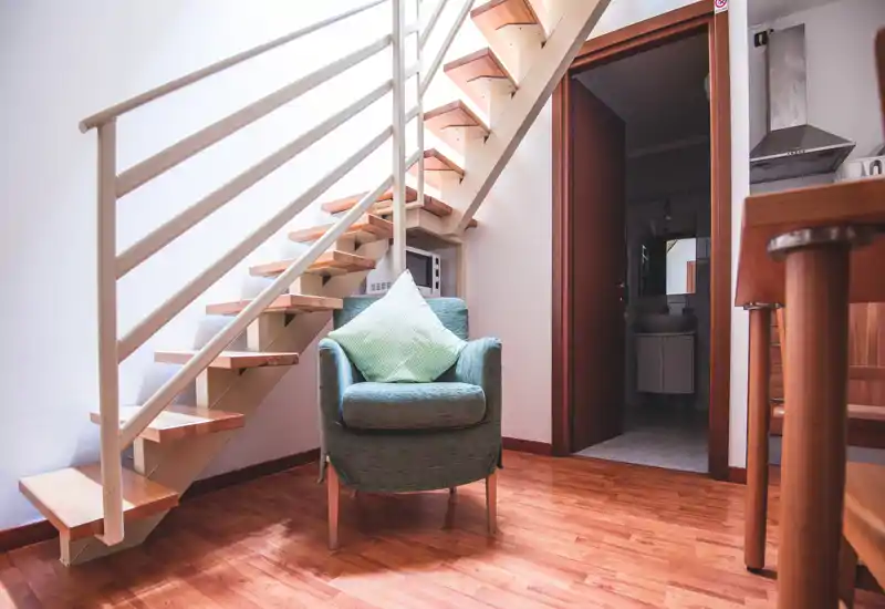 Acogedor rincón de estar bajo una pequeña escalera con un sillón tapizado y cojín decorativo, suelos de madera cálida y luz natural suave — ideal como rincón de lectura en un piso compacto.