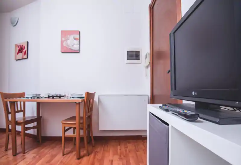 Área de estar/comedor compacta con mesa y sillas de madera, suelos cálidos de parquet, cuadros en la pared y TV sobre mueble blanco: un espacio ordenado y funcional para comer y relajarse.