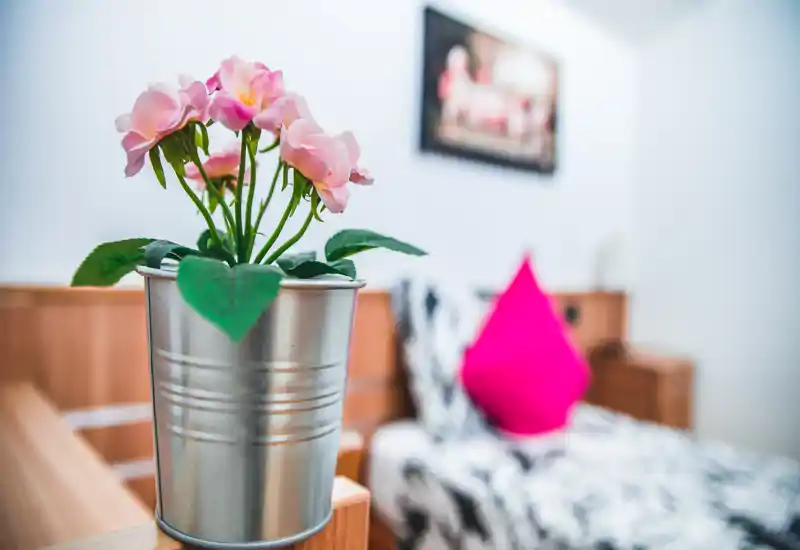 Rincón de dormitorio luminoso y acogedor con cama estampada, cojín rosa intenso y flores decorativas en maceta metálica en primer plano.