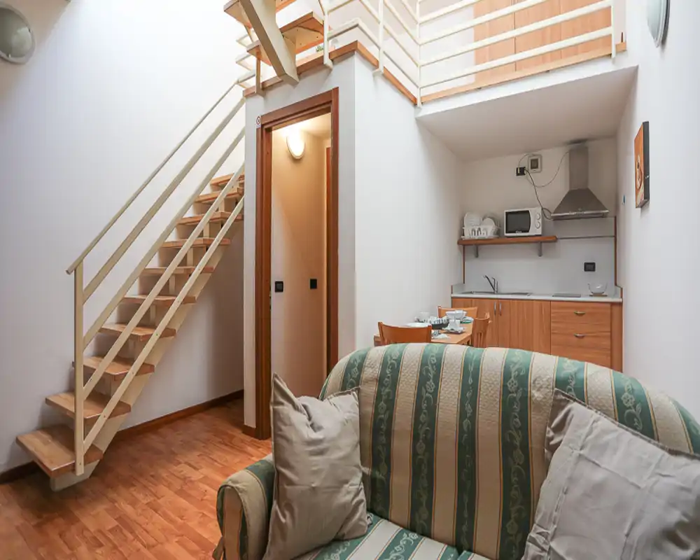 Cozy living area with a patterned sofa, open staircase and a compact kitchenette tucked under a mezzanine — bright, well-staged space ideal for a small apartment.