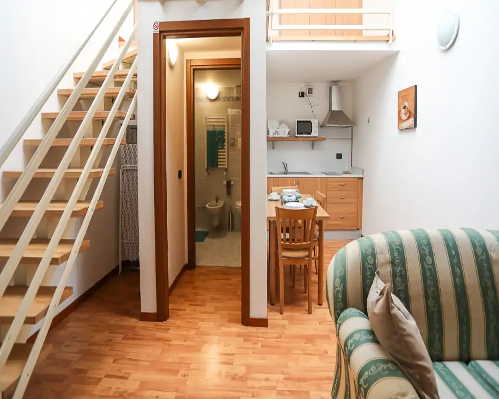 Bright, cozy living area with a striped upholstered sofa, small dining table and an open kitchenette beneath a mezzanine. Wooden floors and a staircase add warmth and vertical interest — a compact, well-lit apartment living space.