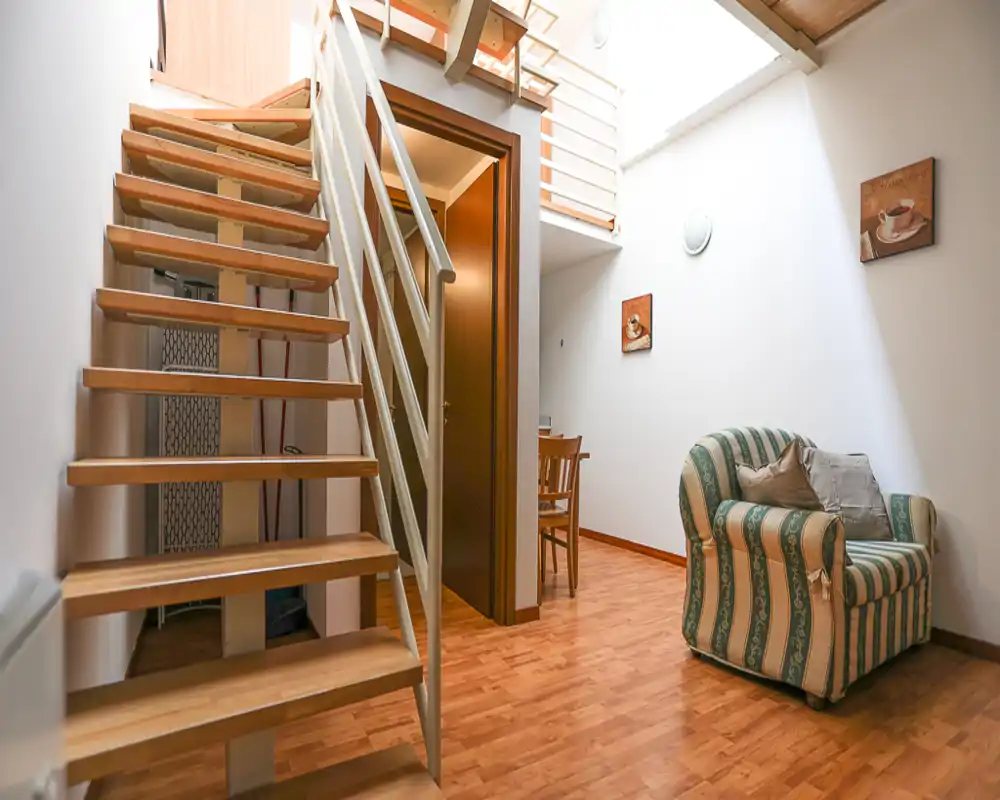 Compact, well-lit living area with a striped armchair, wooden flooring and an open staircase leading to a mezzanine — cozy corner ideal for relaxation.