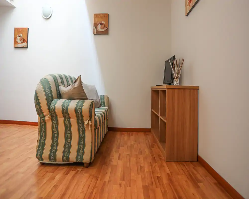 A small, simply furnished living area with a patterned upholstered sofa, wooden floor and a low shelving unit holding a monitor — bright and minimally styled.