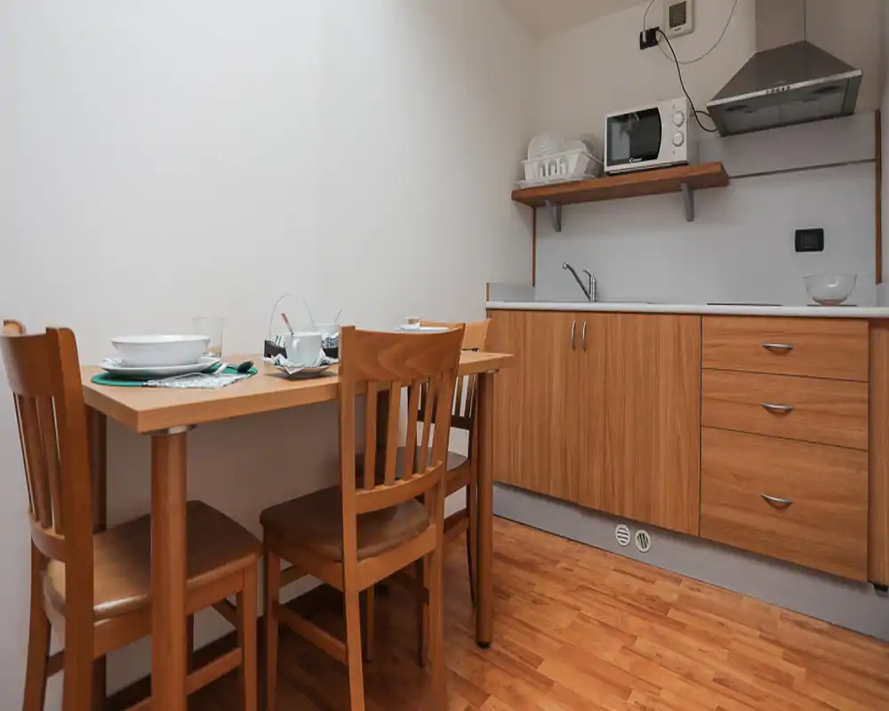 Compact fitted kitchen with wooden cabinets, a small dining table set for two, microwave and extractor hood — warm wooden tones and tidy layout ideal for a short-stay rental.