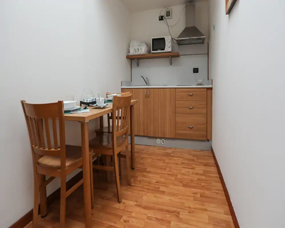 Compact kitchenette with a small wooden dining table and two chairs. The fitted cabinets, sink and extractor hood create a practical cooking area in a bright, tidy space.