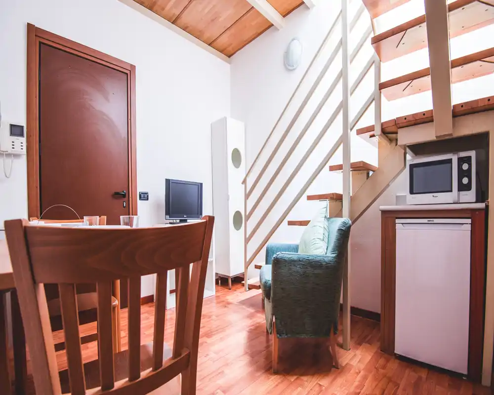 Área de estar compacta y luminosa con un sillón cómodo, mesa pequeña y televisor — suelos de madera y cocina bajo la escalera crean un ambiente acogedor.